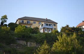 Ein gelbes Gebäude mit der Aufschrift 'Donaublick' auf einem Hügel, umgeben von Bäumen und blauem Himmel.