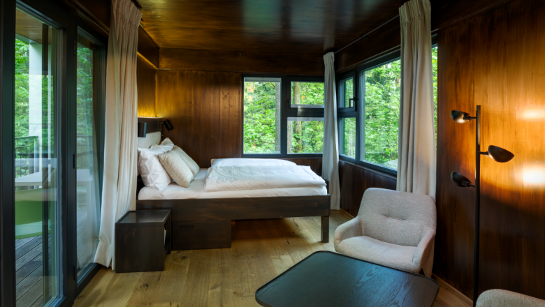 Innenansicht eines modernen Baumhauses mit Holzwänden, Bett, Sessel und großen Fenstern mit Blick ins Grüne.