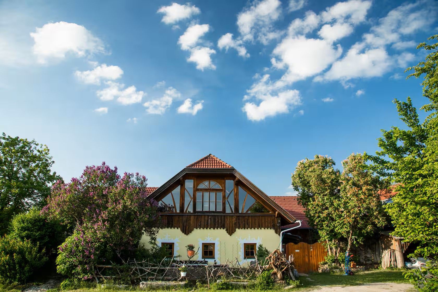 Haus mit rotem Ziegeldach, umgeben von Bäumen und Sträuchern unter blauem Himmel mit Wolken.
