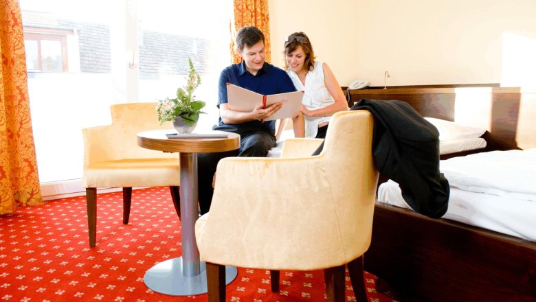 Ein Paar sitzt in einem Hotelzimmer auf einem Bett und schaut in ein Buch. Der Raum hat rote Teppiche und gelbe Vorhänge.