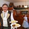 Ein Mann in traditioneller Kleidung serviert Schweinsbraten mit Knödeln in einem rustikalen Gasthaus. Im Hintergrund ein Mann und ein Mädchen im Kleid.
