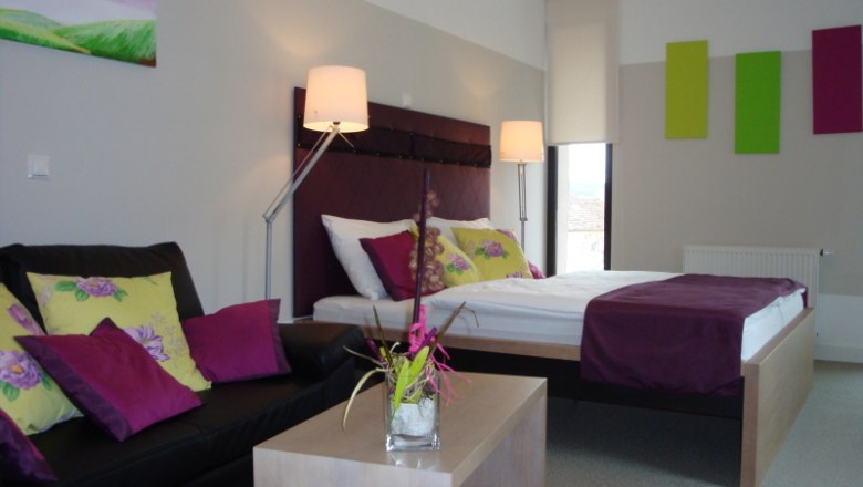 Modern eingerichtetes Hotelzimmer mit Doppelbett, Sofa und Dekoration in Lila- und Grüntönen.