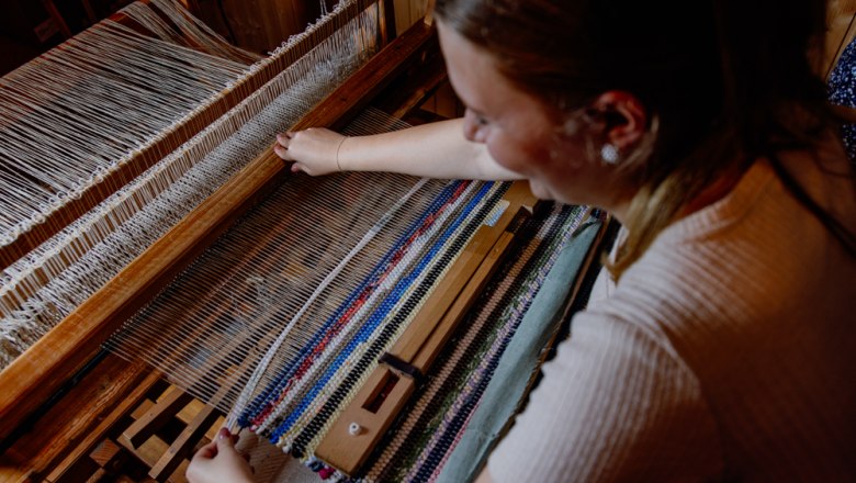 Eine Frau webt an einem Webstuhl einen bunten Fleckerlteppich.