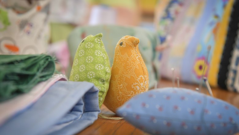 Zwei Stoffvögel, einer grün und einer orange, auf einem Tisch mit Stoffen und Nähutensilien.