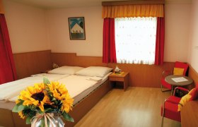 Ein gemütliches Zimmer mit Doppelbett, roten Vorhängen, einem Tisch mit Stühlen und einem Sonnenblumenstrauß im Vordergrund.