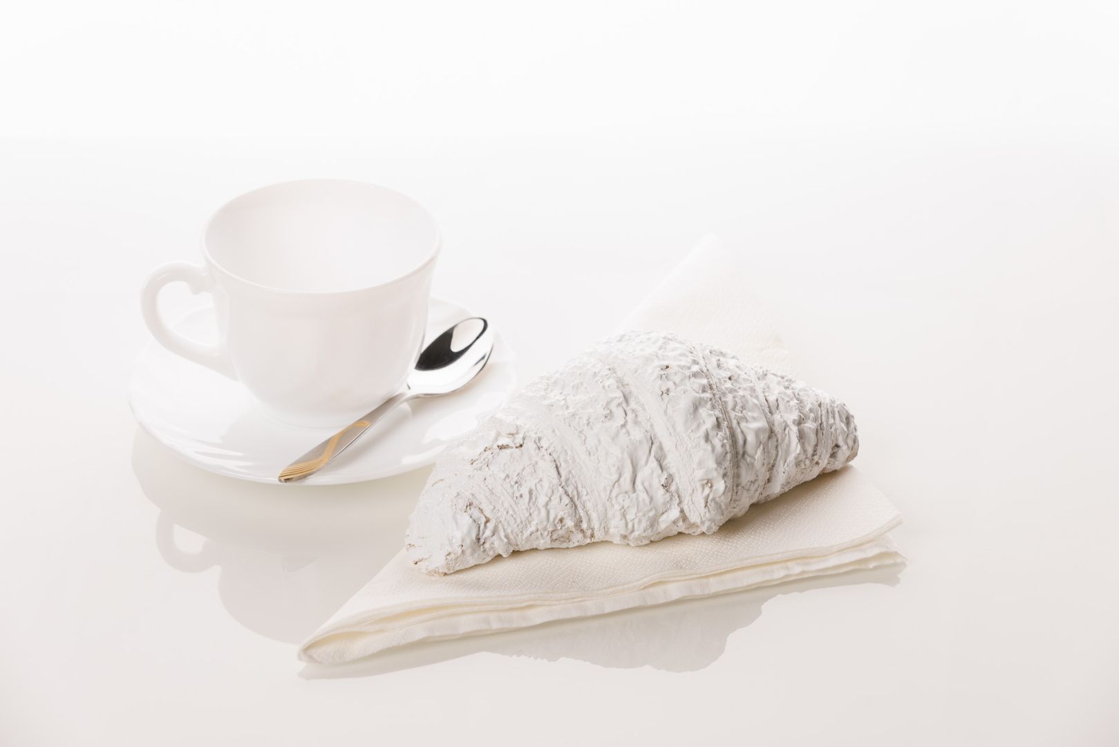 Luxury Life - Weiße Tasse mit einem Löffel am Rand der Untertasse, daneben liegt ein Croissant, das 