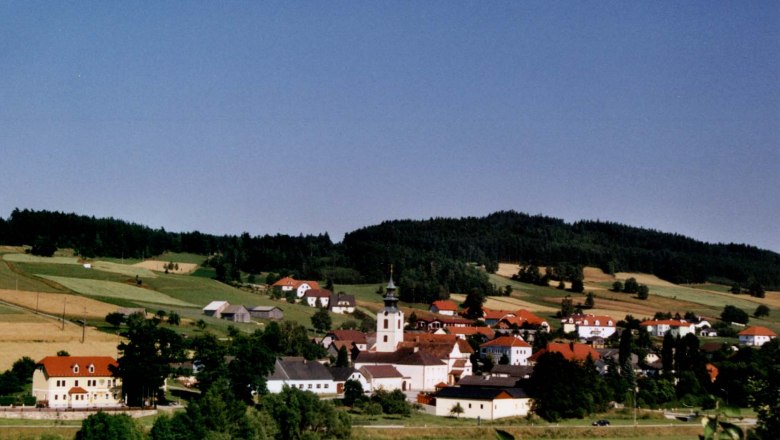 Panorama eines Dorfes mit Kirche und roten Dächern, umgeben von grünen Feldern und Wäldern.