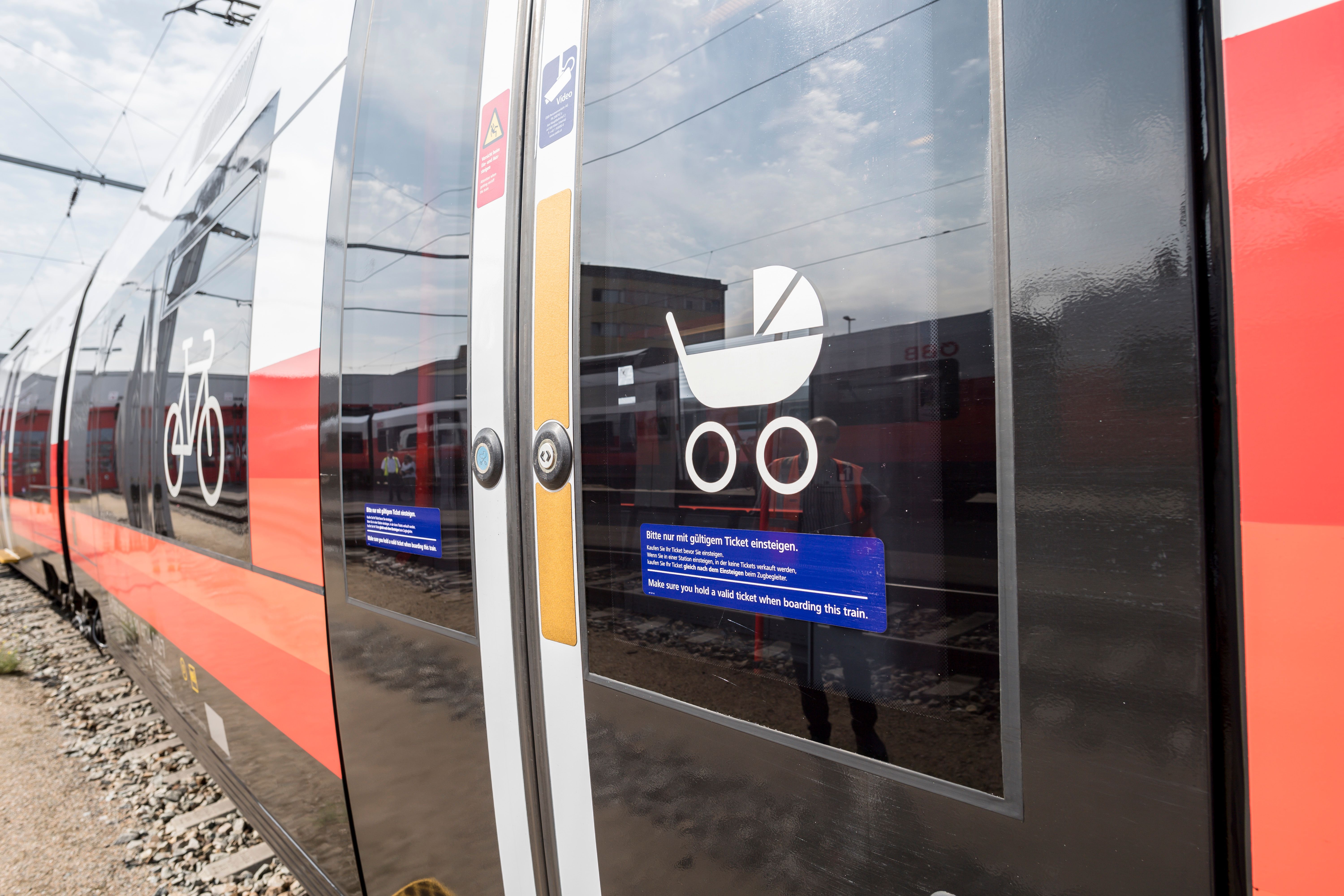 Nahaufnahme einer Zugtür mit Symbolen für Fahrrad und Kinderwagen. Im Hintergrund sind weitere Züge und ein Gebäude zu sehen.