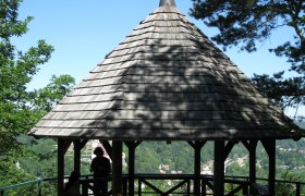 Ein hölzerner Pavillon mit spitzem Dach auf einem Hügel, umgeben von Bäumen, mit Blick auf eine grüne Landschaft.