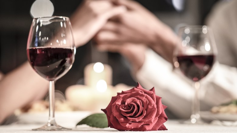 Zwei Weingläser, eine rote Rose und ein Paar, das sich die Hände hält, bei Kerzenschein.