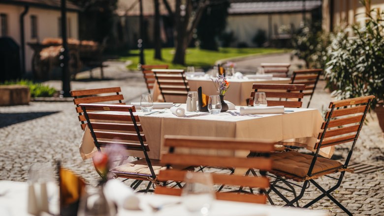 Ein sonniger Gastgarten mit Holztischen und -stühlen, gedeckt mit weißen Tischdecken und Gläsern, in einem gepflasterten Innenhof.