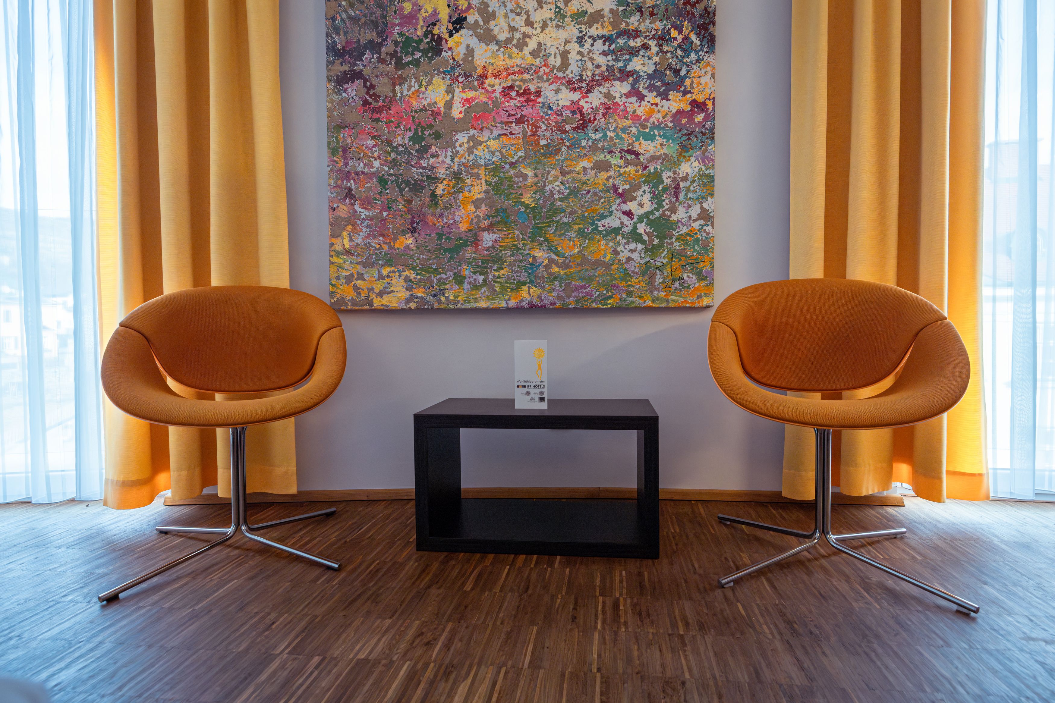Zwei orangefarbene Stühle vor einem bunten Gemälde und gelben Vorhängen.