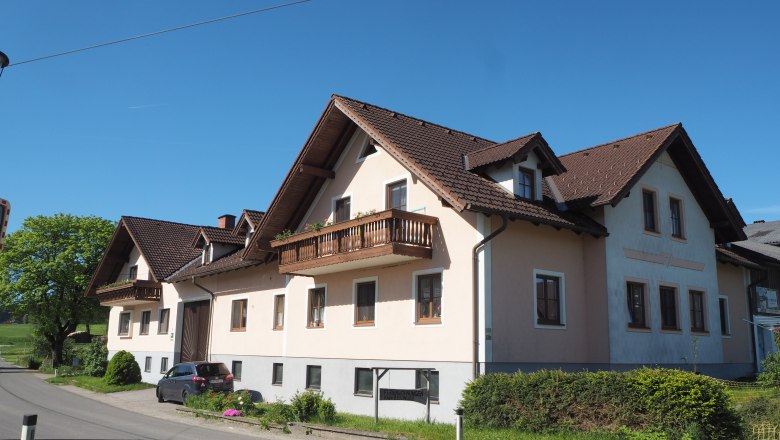 Ein großes Bauernhaus mit braunem Dach und Balkonen an einer Landstraße bei klarem Himmel.