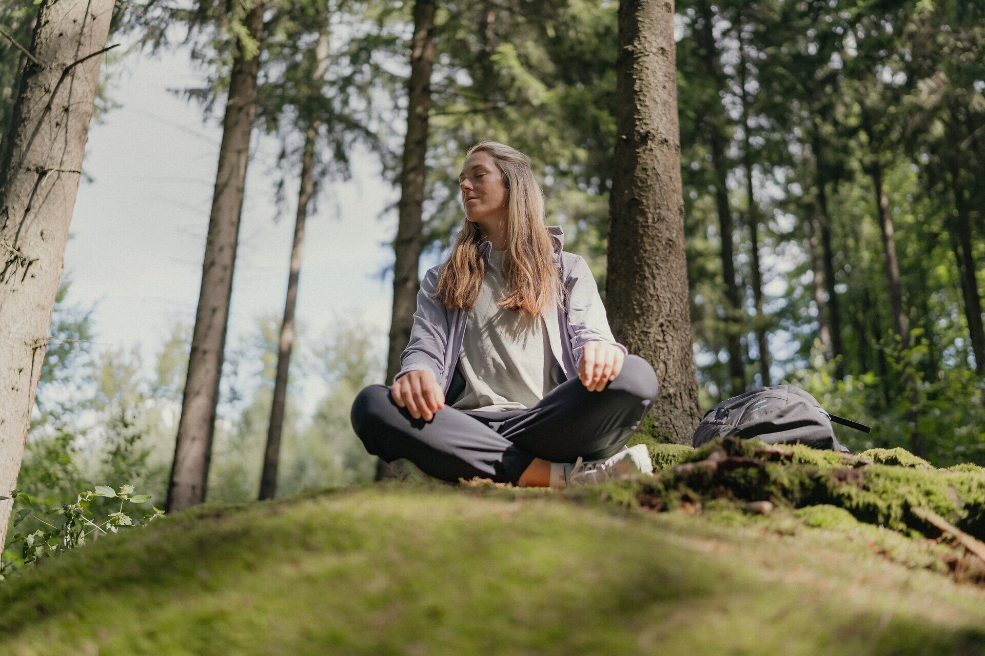 Frau sitzt im Schneidersitz im lichtdurchfluteten Wald, der Kopf zur Seite geneigt und die Augen geschlossen. Neben ihr liegt ein Rucksack, während sie die frische Luft und Ruhe genießt.