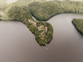 Drohnenaufnahme der bewaldeten Halbinsel mit der Ruine Dobra im Stausee Dobra; die Ruine liegt von Wasser umgeben, mit kleinen Booten und Wiesenfl&auml;chen am Ufer.