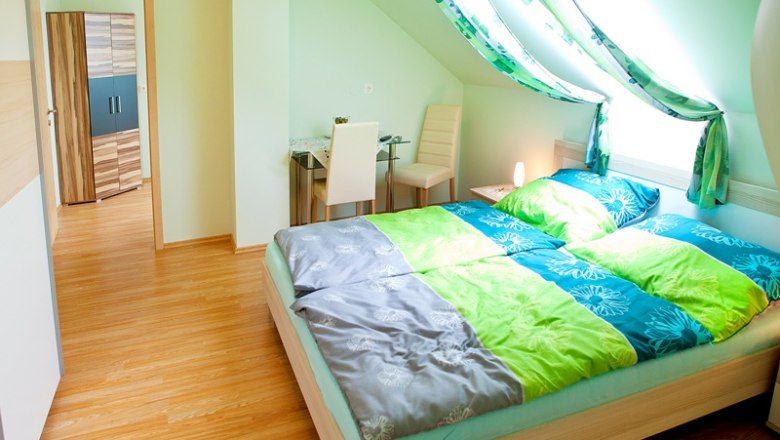 Helles Schlafzimmer mit Dachschräge, Doppelbett, Schreibtisch und Stuhl.