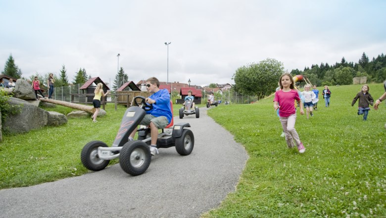 Kinder spielen auf einem Erlebnisspielplatz mit Kettcars und laufen &uuml;ber eine Wiese.