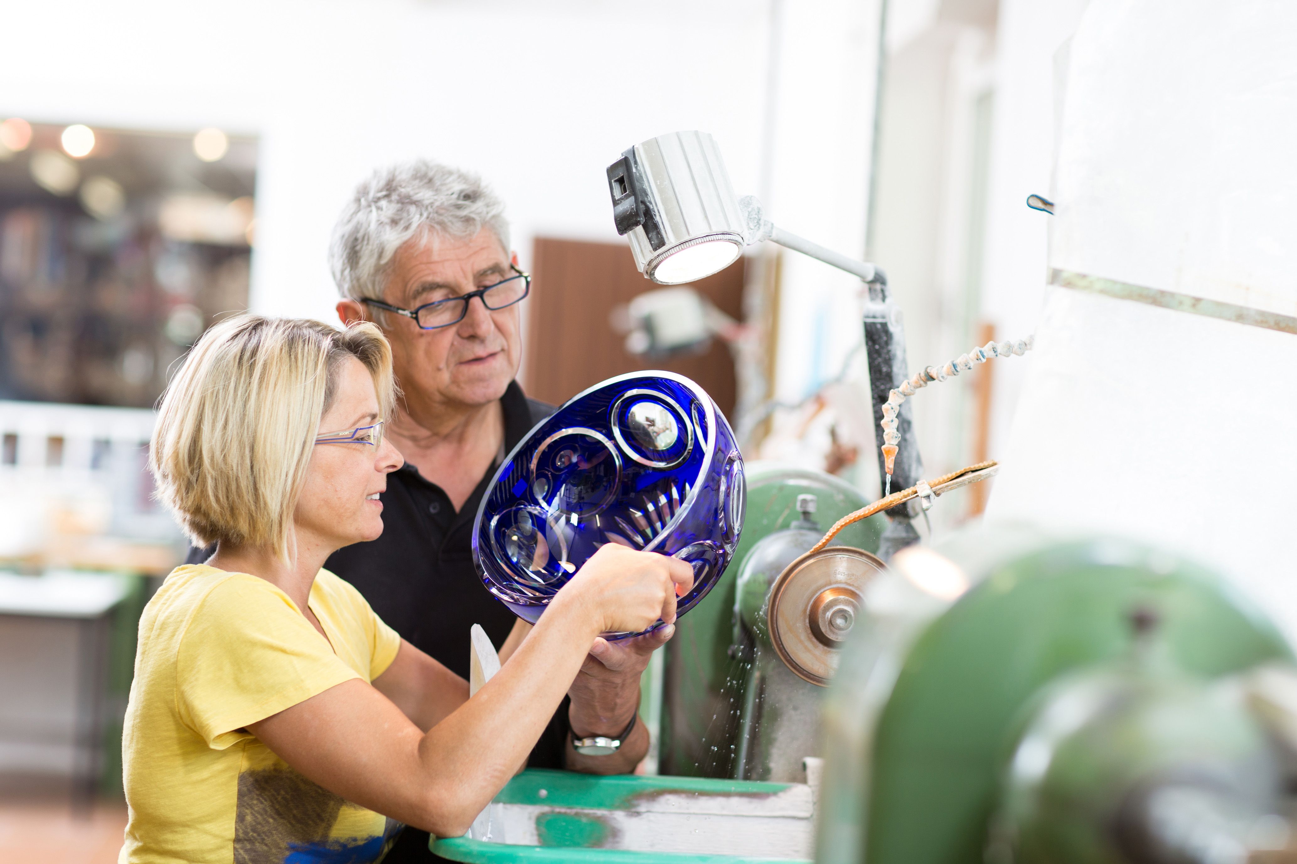 Zwei Personen arbeiten an einer Glasschleifmaschine mit einer blauen Glasschale.