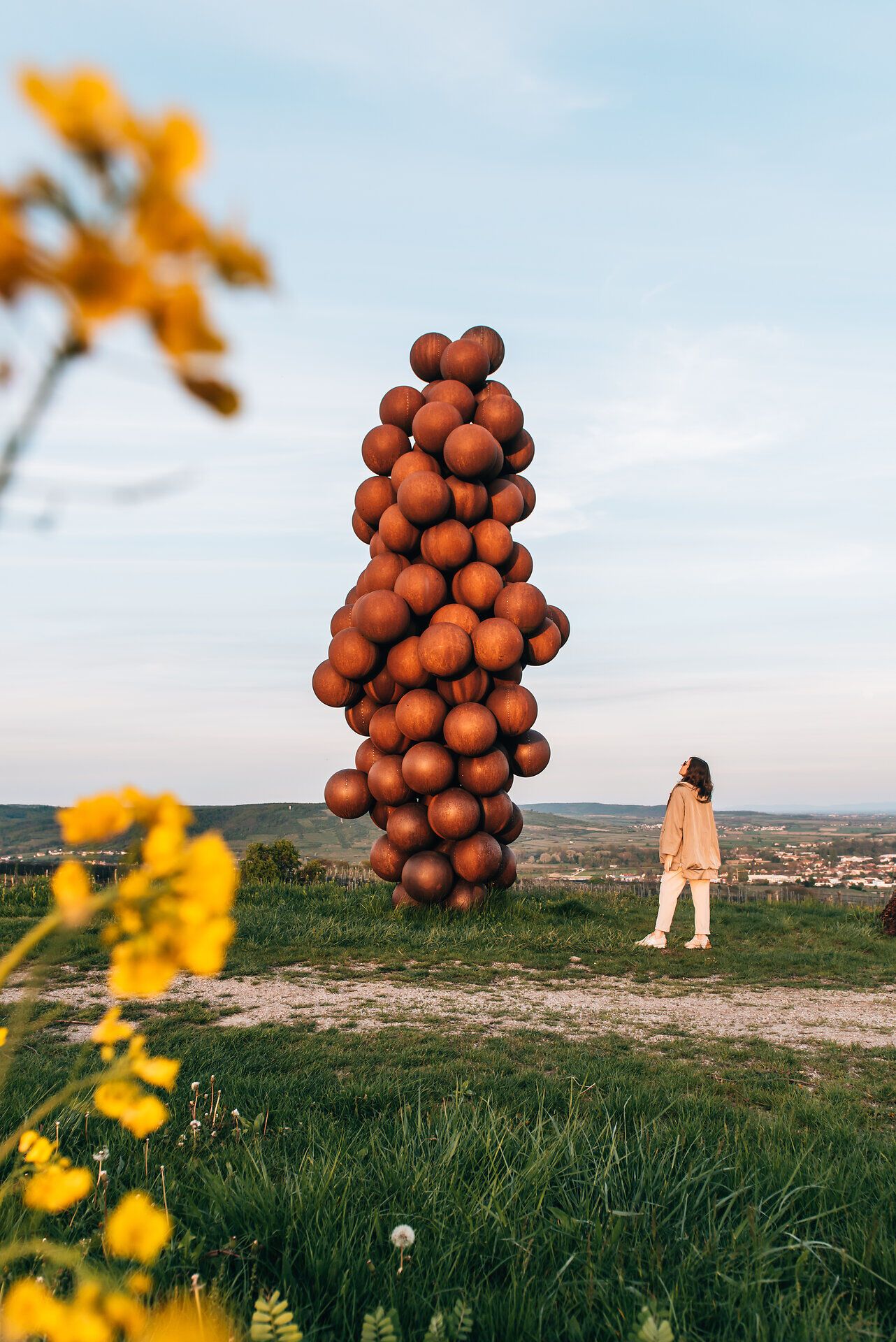 Eine beeindruckende Skulptur aus zahlreichen, rustikalen Kugeln erhebt sich majestätisch in die Höhe und spiegelt die kreative Verbindung zur Weinregion wider. Umgeben von sanften Hügeln und blühenden Wildblumen, lädt dieser Ort dazu ein, die Schönheit der Natur und die Kunst in harmonischem Einklang zu genießen.