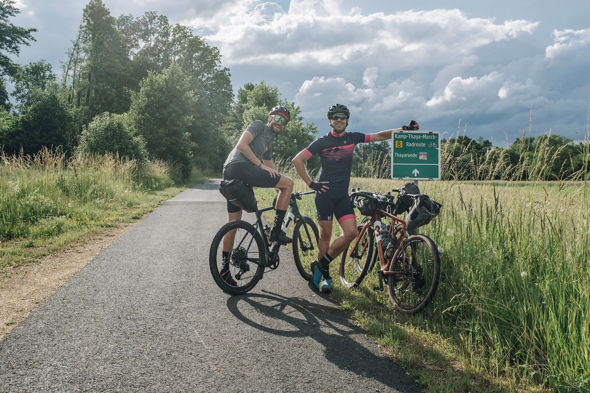 Zwei Radfahrer genießen die frische Luft und die malerische Landschaft entlang der Kamp-Thaya-March Radroute. Umgeben von üppigem Grün und sanften Hügeln, strahlt die Szenerie eine einladende Ruhe aus, die zum Verweilen einlädt.