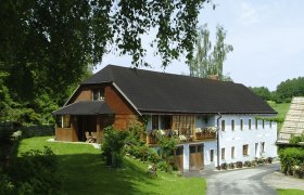 Ein traditionelles Bauernhaus mit Blumen auf dem Balkon, umgeben von grüner Landschaft und Bäumen.