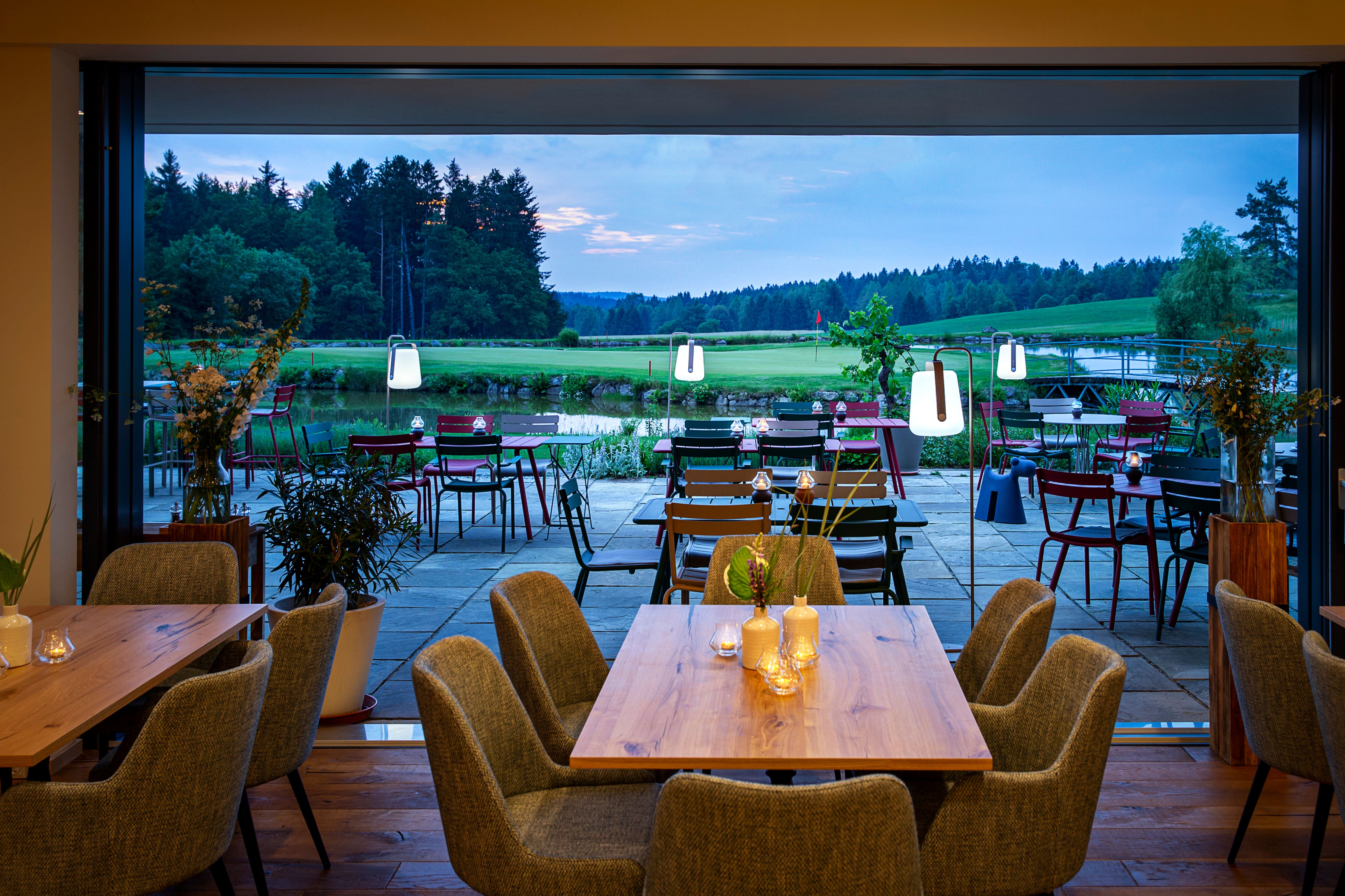 Blick aus dem Restaurant des Golfresorts Haugschlag auf die Terrasse, den Golfplatz mit Wasserhindernis und den Wald bei Abendlicht.