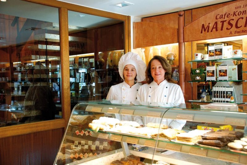 Zwei Frauen in Konditoruniformen stehen hinter einer Theke mit Gebäck in einer Bäckerei.