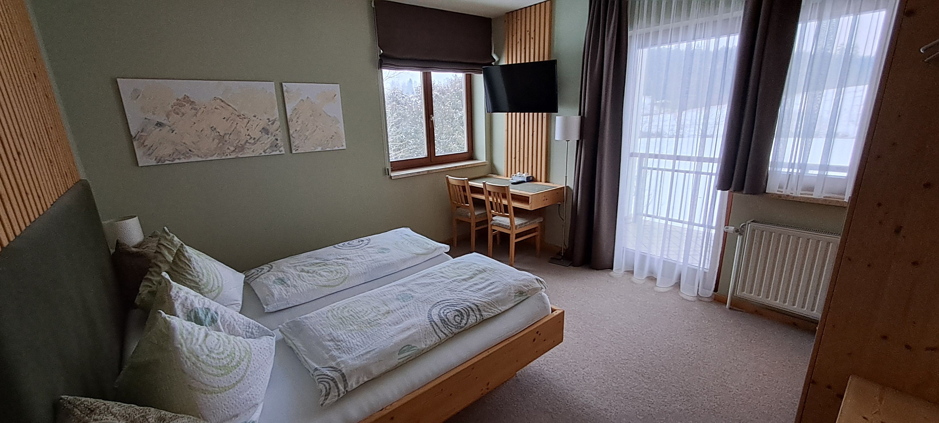 Helles Doppelzimmer, Schreibtisch, Fernseher und Balkonblick auf verschneite Bäume.