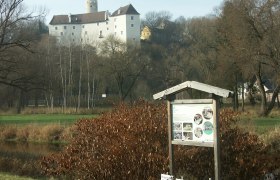 Ein Lehrpfad-Schild vor einer Burg in einer ländlichen Landschaft.