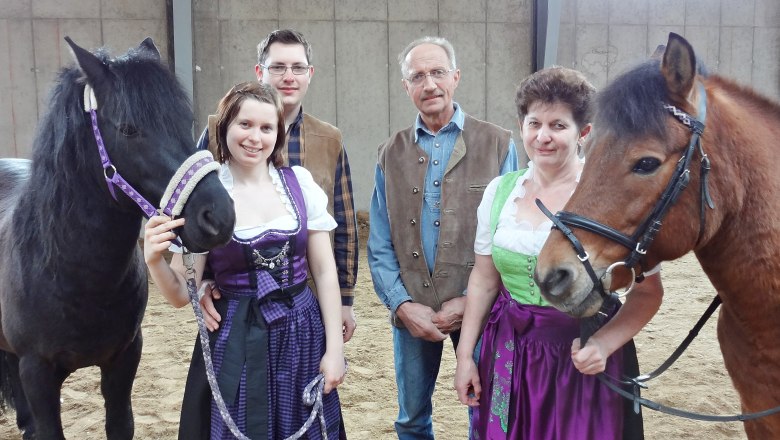 A family in traditional dress stands with two horses in an indoor riding arena.