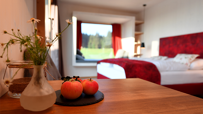 A modern hotel room with a red bedspread, a table with apples and a window with a view of the greenery.