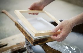 Nahaufnahme von Händen, die einen Holzrahmen über einer Papiermasse in einer traditionellen Papiermühle halten.