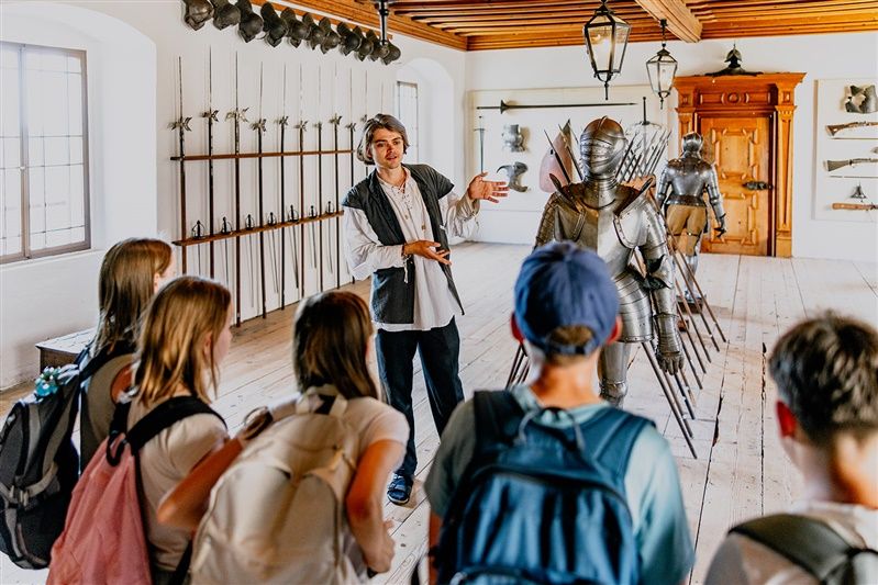 Ein Führer erklärt einer Gruppe von Jugendlichen in einem Raum mit mittelalterlichen Rüstungen und Waffen die Ausstellung im Renaissanceschloss Rosenburg.