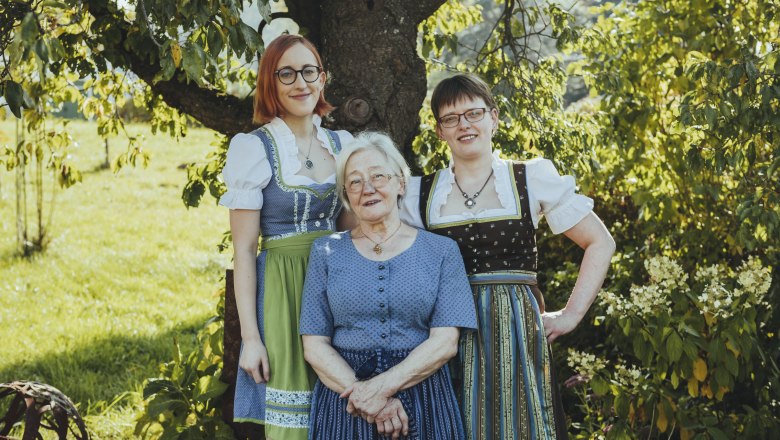 Drei Frauen in traditioneller Tracht stehen lächelnd unter einem Baum im Freien.