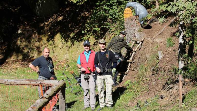 Drei Männer mit Bögen im Wald, ein Mann klettert eine Leiter hoch.