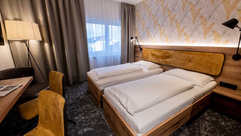 Modernes G&auml;stezimmer mit zwei Einzelbetten, Holzm&ouml;beln, gelben St&uuml;hlen und einer Stehlampe. Helle Vorh&auml;nge und gemusterte Tapete an der Wand.