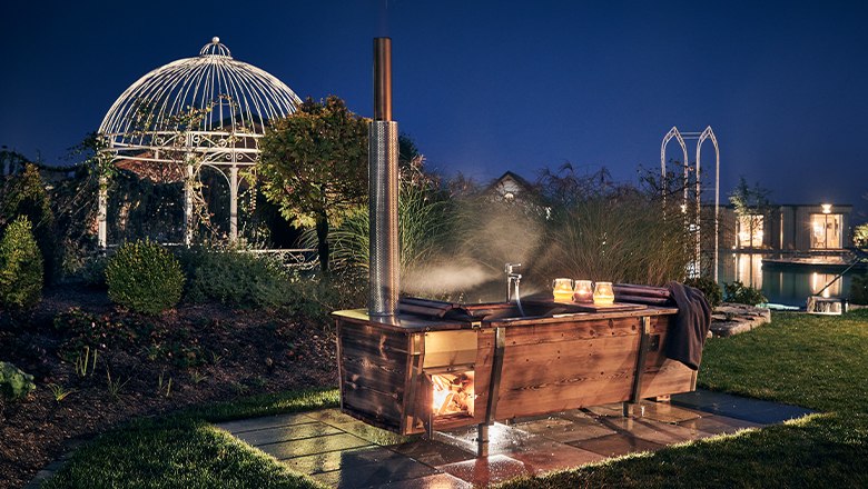 Eine Holzbadewanne mit Feuerheizung im Freien, umgeben von Garten und Pavillon bei Nacht.
