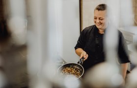 A cook in a black uniform holds a pan of roasted vegetables and smiles.