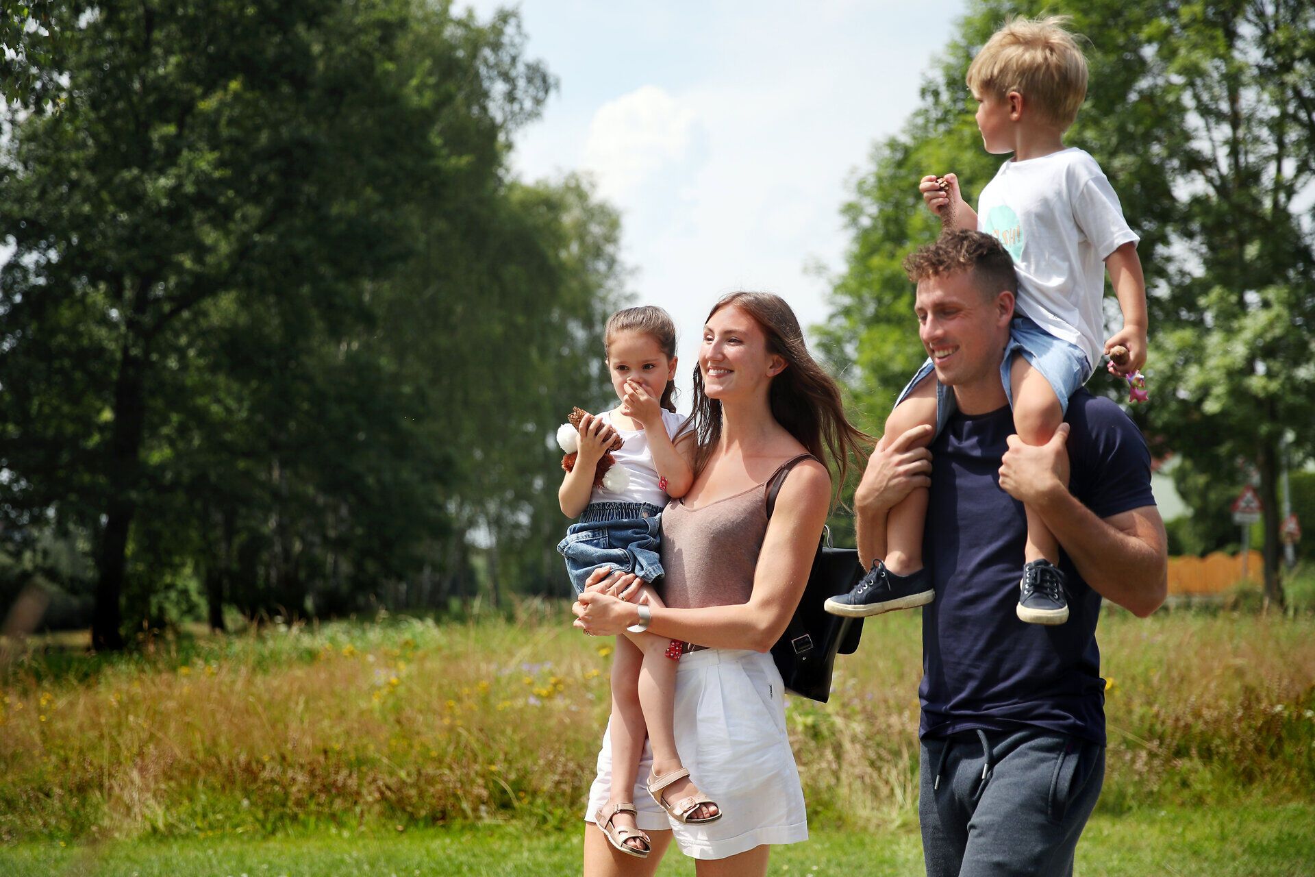 Eine fröhliche Familie genießt einen sonnigen Tag im Freien, während die Kinder voller Freude auf den Schultern ihres Vaters sitzen. Umgeben von der üppigen Natur und dem sanften Licht, strahlt die Szene eine herzliche und unbeschwerte Atmosphäre aus.
