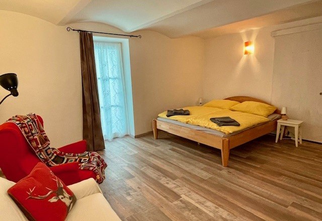 Gemütliches Schlafzimmer mit Holzboden, Doppelbett, rotem Sessel und Fenster mit Vorhang.