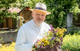 Ein Mann in weißem Gewand und Strohhut hält einen Korb mit bunten Kräutern in einem Garten.