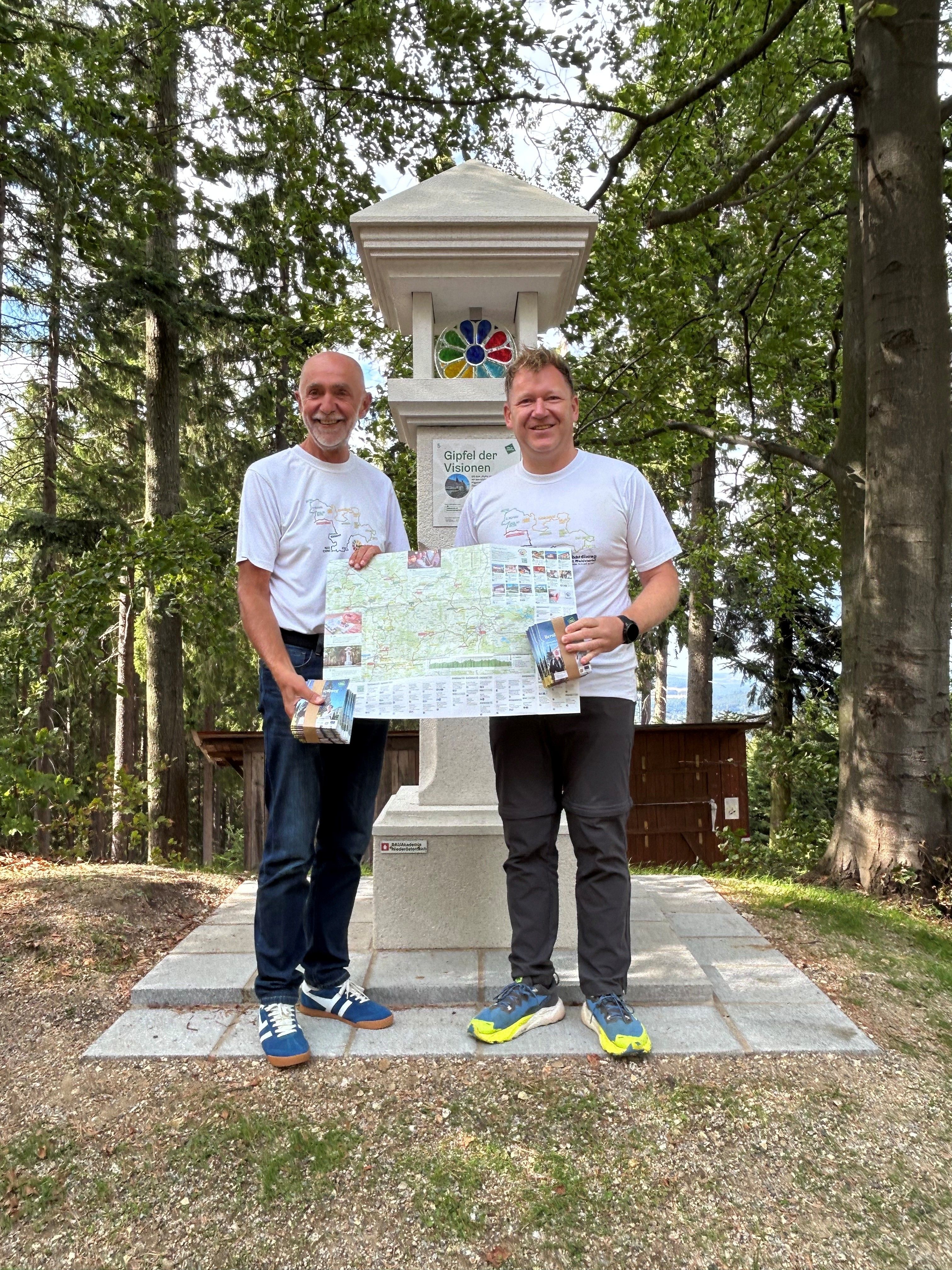 Zwei Männer halten die neue Wanderkarte vom Bernhardiweg geöffnet in den Händen und präsentieren sie. Sie stehen vor einem Marterl im Wald am Johannesberg, der eine Station am Bernhardiweg ist.