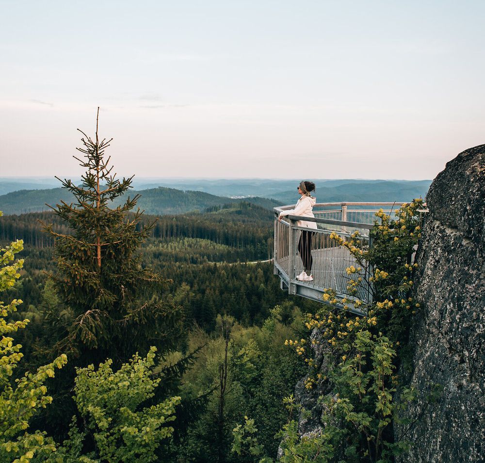 Ein atemberaubender Ausblick eröffnet sich über die sanften Hügel und dichten Wälder, während die Abendsonne den Himmel in sanften Pastelltönen färbt. Die frische Bergluft und das sanfte Rascheln der Blätter laden dazu ein, einen Moment innezuhalten und die Schönheit der Natur zu genießen.