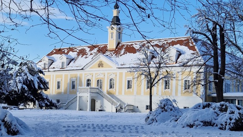 Schloss Haindorf im Winter mit Schnee bedeckt, blauer Himmel und kahle Bäume im Vordergrund.