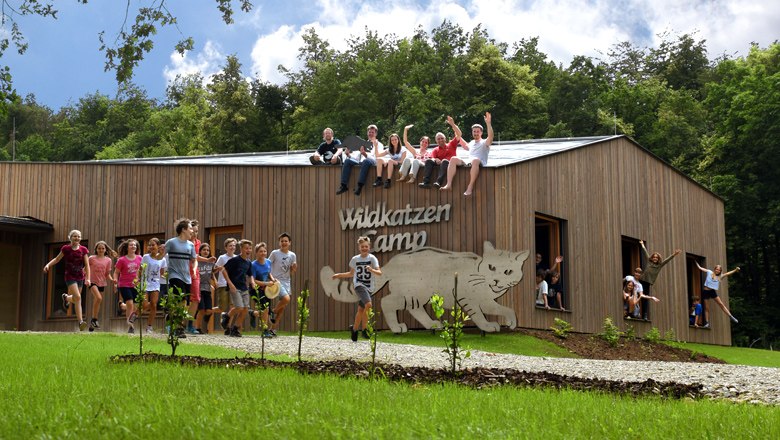 Eine Gruppe von Kindern und Erwachsenen sitzt auf einem Holzgebäude mit der Aufschrift 'Wildkatzen Camp'. Einige Kinder stehen und springen davor.