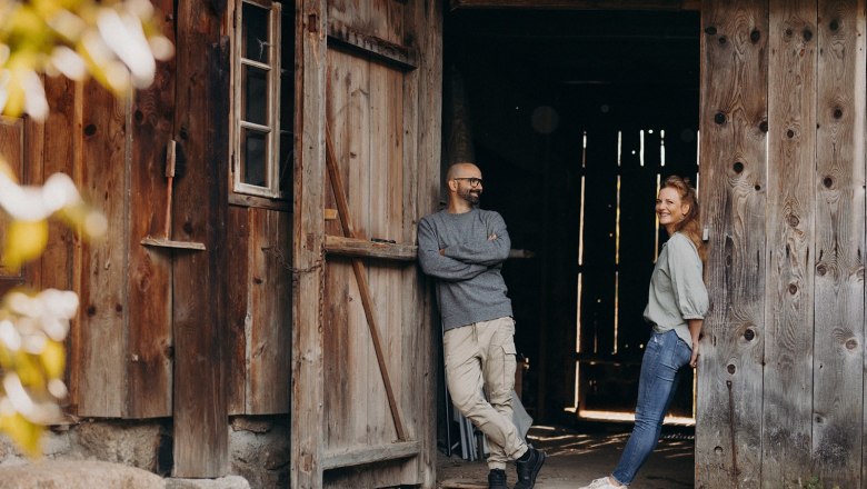 Zwei Personen stehen lächelnd vor einer alten Holzhütte.