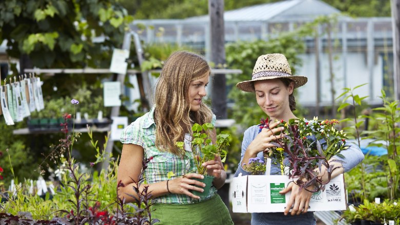 Zwei Frauen in einem Gartenladen, eine mit Strohhut, halten Pflanzen in der Hand.