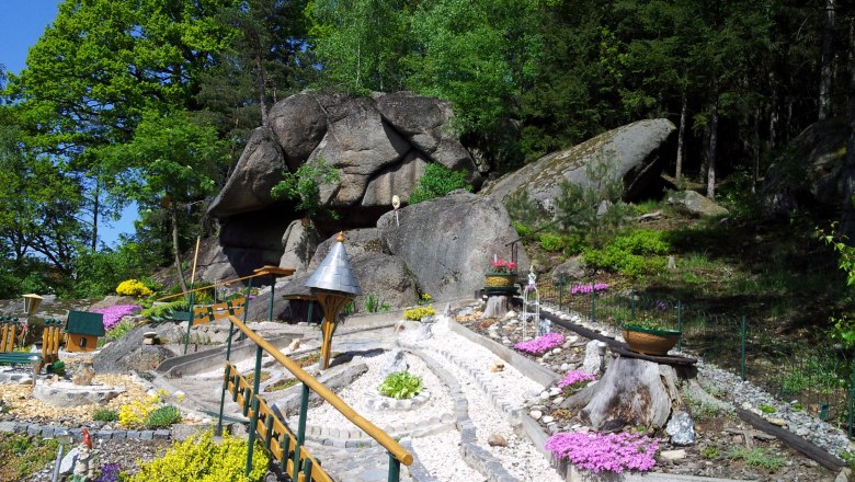 Ein gepflegter Garten mit Steinen, Blumen und einem kleinen Weg vor großen Felsen und Bäumen.