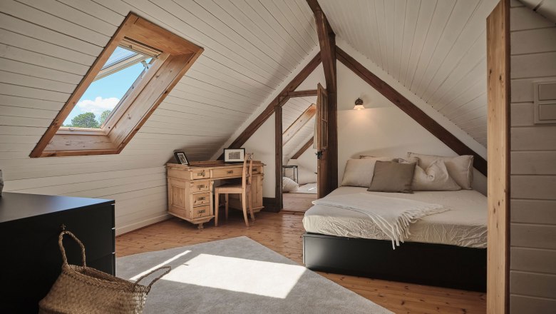 Gemütliches Dachzimmer mit Holzbalken, Bett, Schreibtisch und Dachfenster.