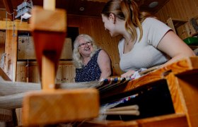 Zwei Frauen beim Weben eines Fleckerlteppichs in einem holzgetäfelten Raum.